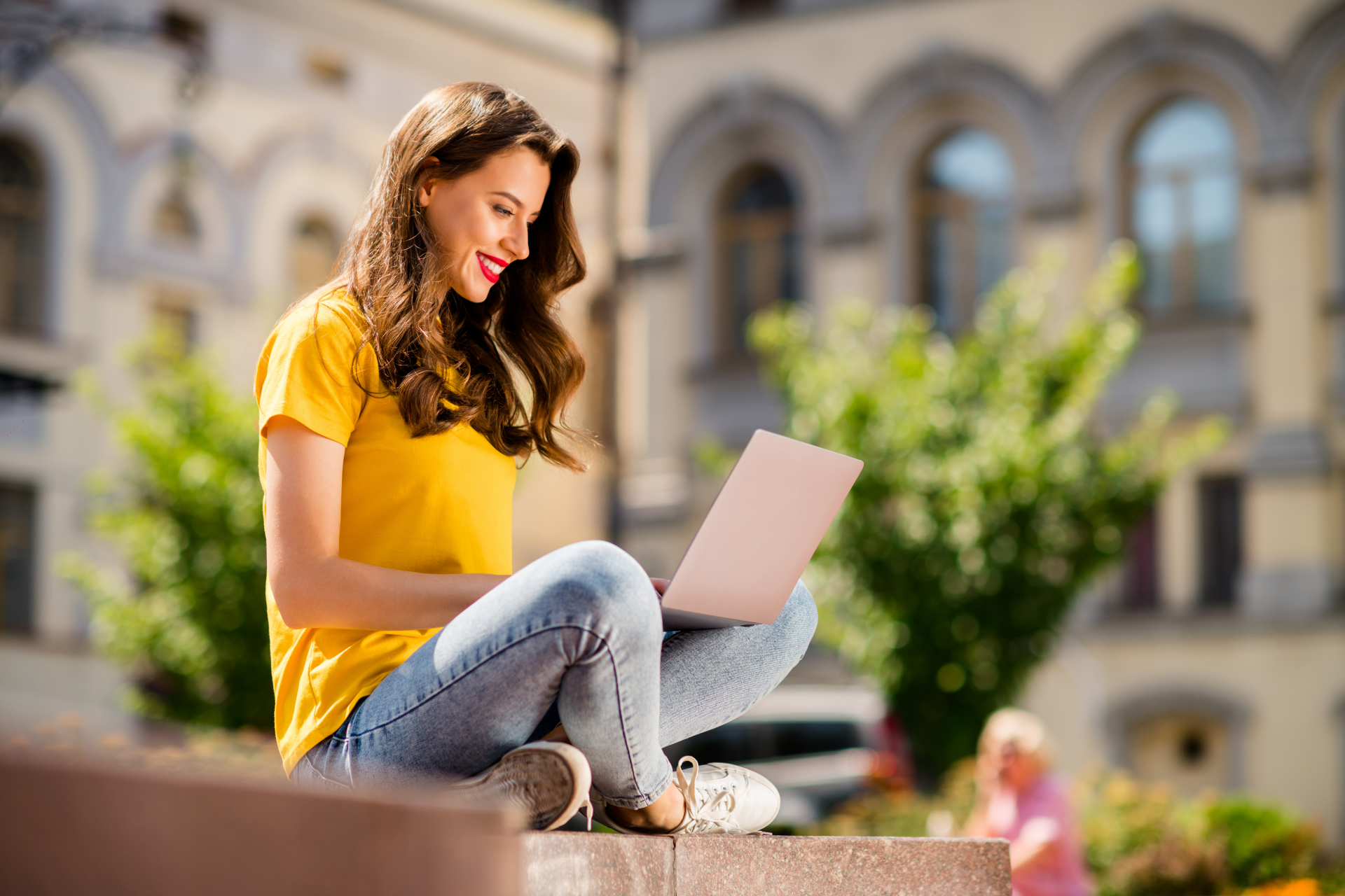 Fiatal lány ül egy lépcsőn a napsütésben laptoppal az ölében háttérben egy kollegium épülete